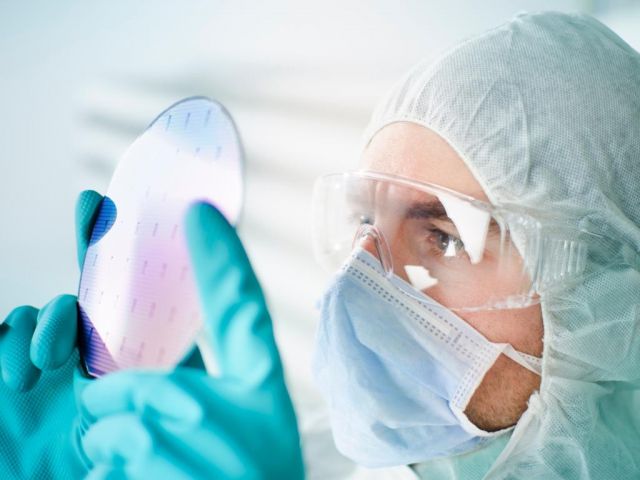 A technical employee in protective clothing for a clean room optically examines a silicon wafer