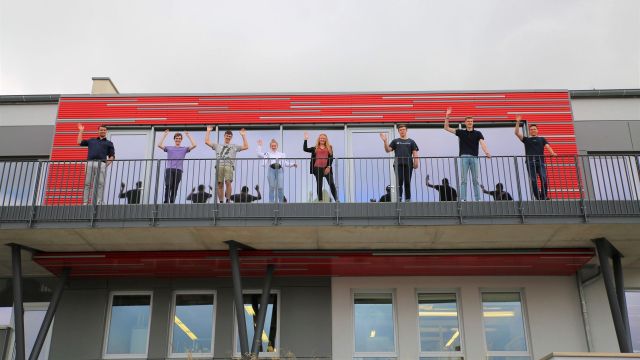 Neue Studierende und Auszubildende stehen auf dem Balkon des Micro-Hybrid Firmengebäudes und winken